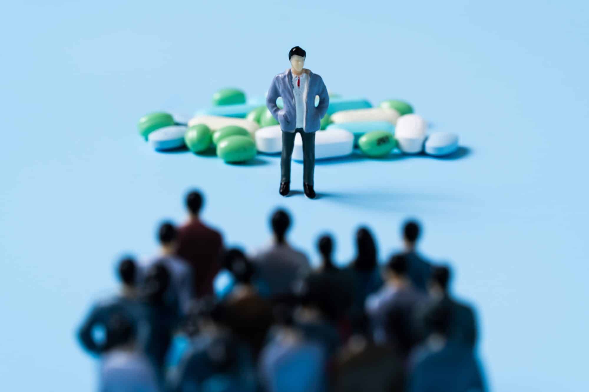 A miniature figure of a man stands in front of a pile of medicine pills, with a crowd of miniature people crowded in the forefront, facing the individual guarding the medicine.