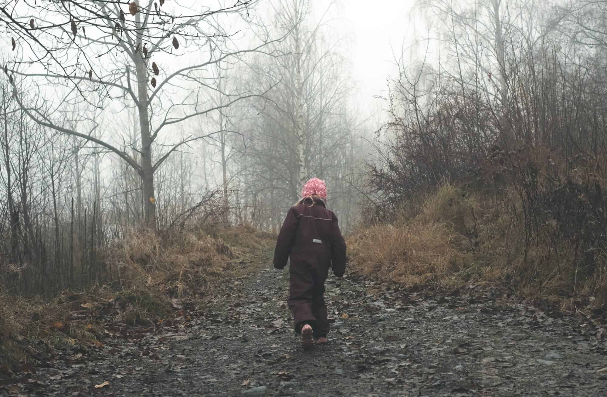 A child in a snow suit walks down a foggy forest trail in the winter.