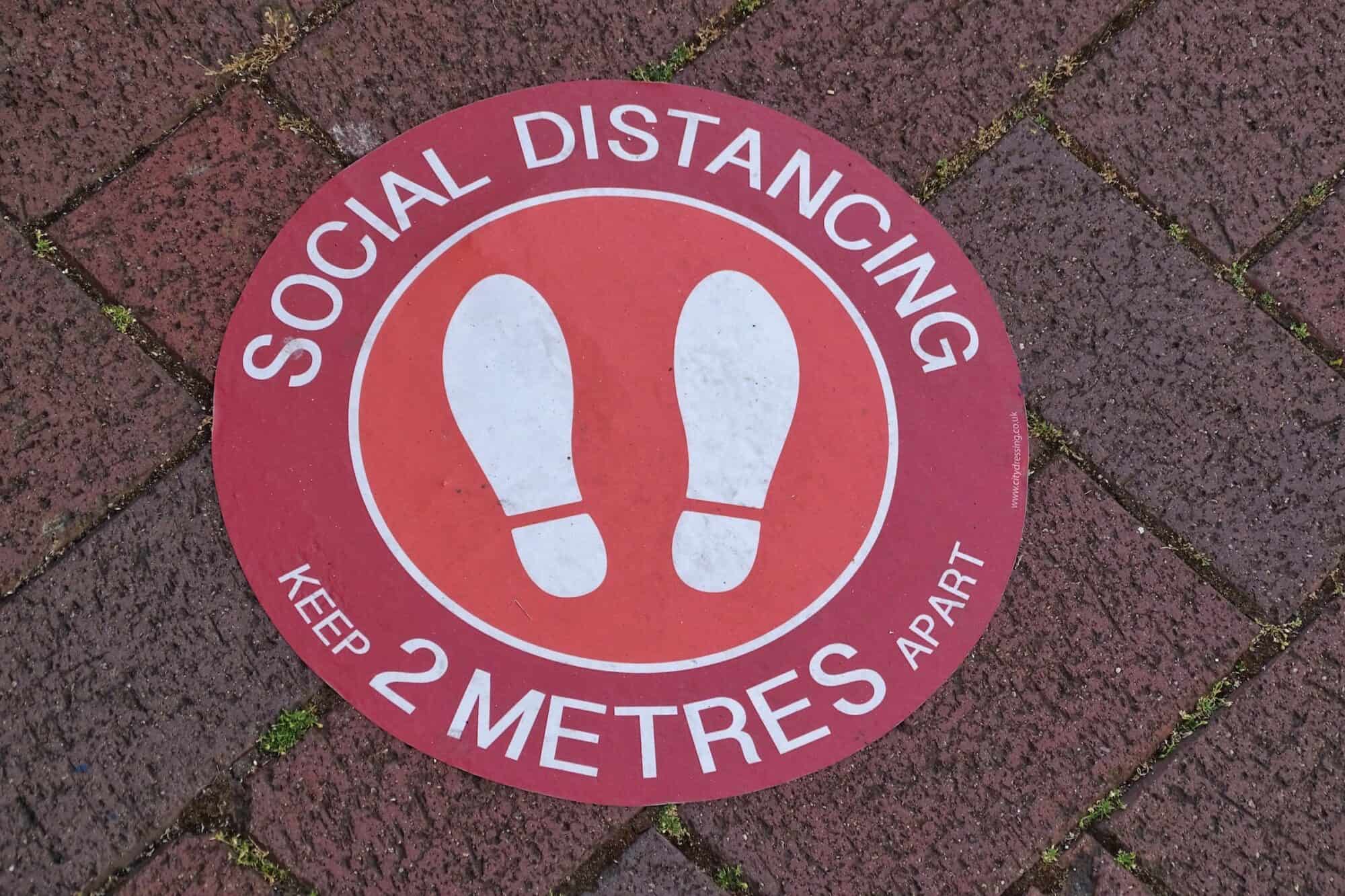 A temporary on the sign placed on a paved floor reads "Social Distancing: keep 2 metres apart".