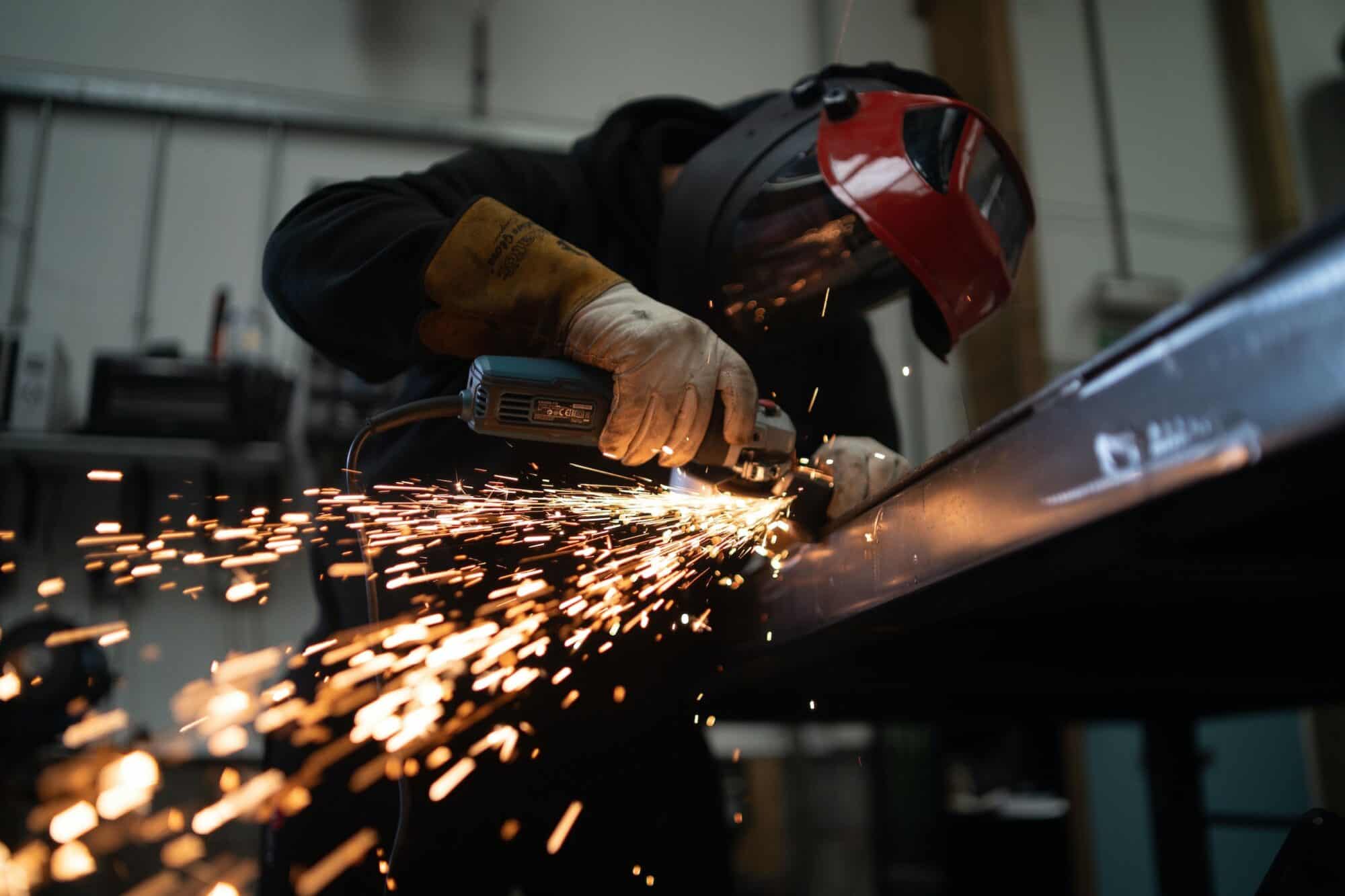 A worker assembles metal in a factory amid sparks emanating from their tool.