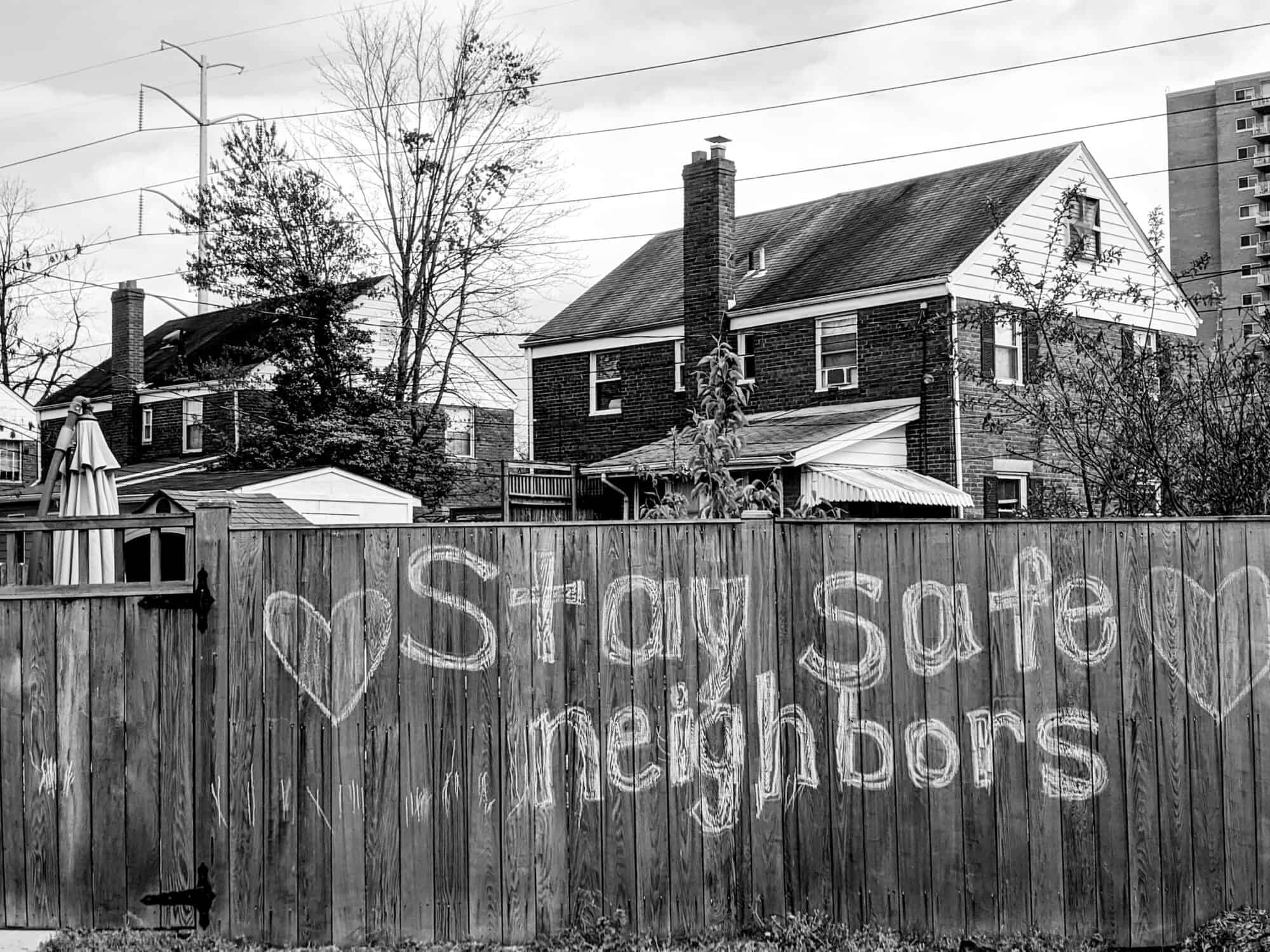 A photo of a neighbourhood fence with "Stay safe neighbours" drawn in chalk behind housing.