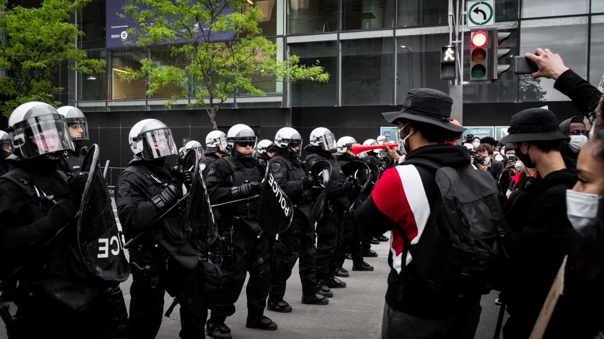 A line of police dressed in riot gear confront protestors on a city street.