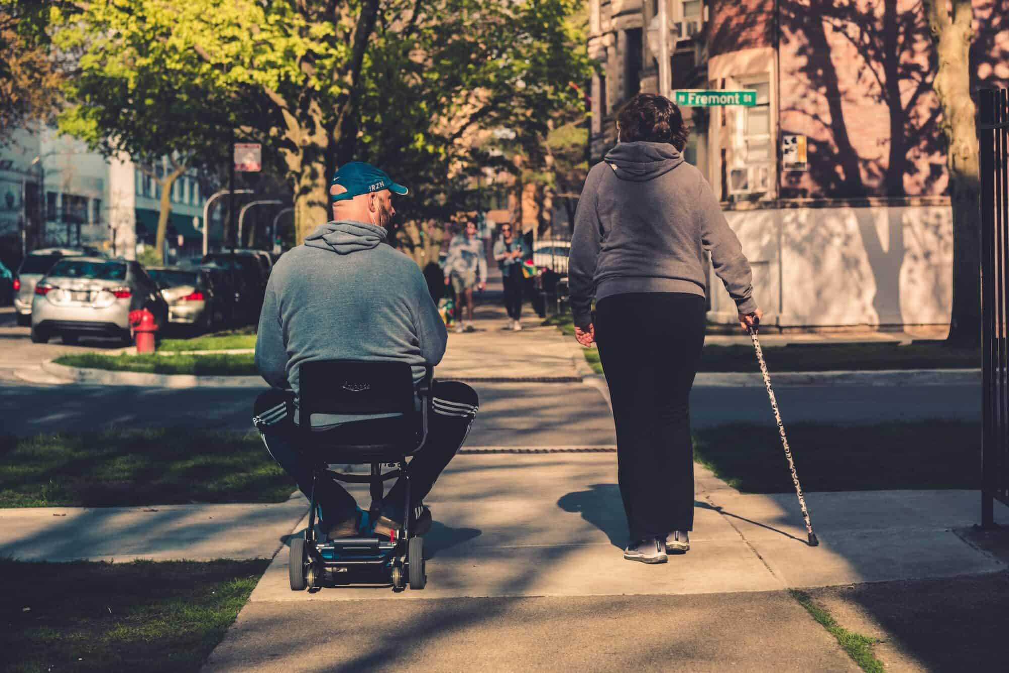 Man in gray hoodie and black pants sitting on black and gray mobility device on a city sidewalk text to elderly woman using a cane to assist in walking.