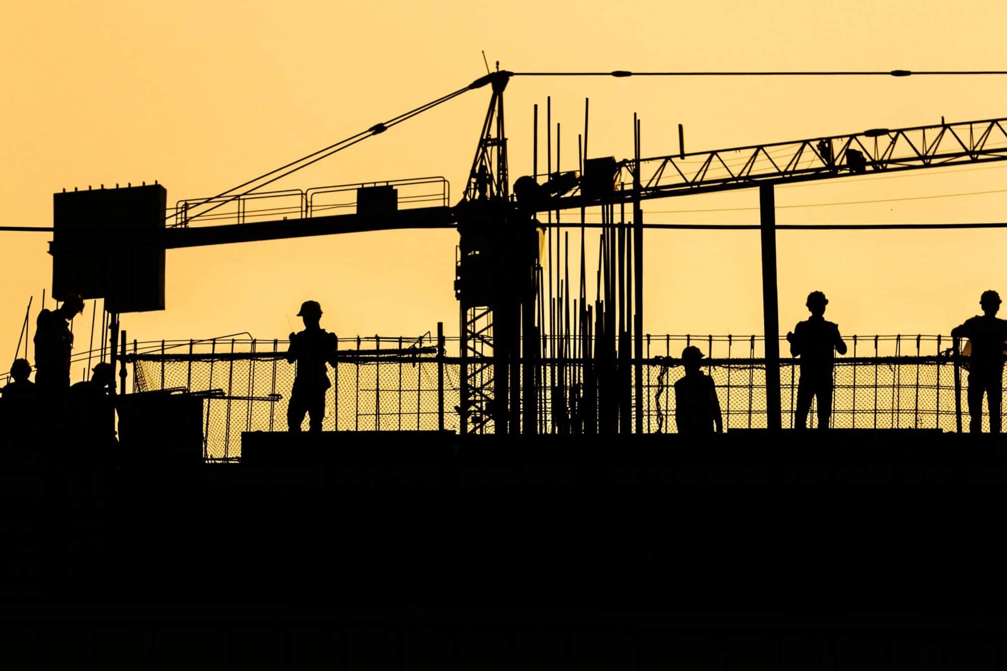 Silhouettes of construction workers on top of a project in front of a sunset sky.