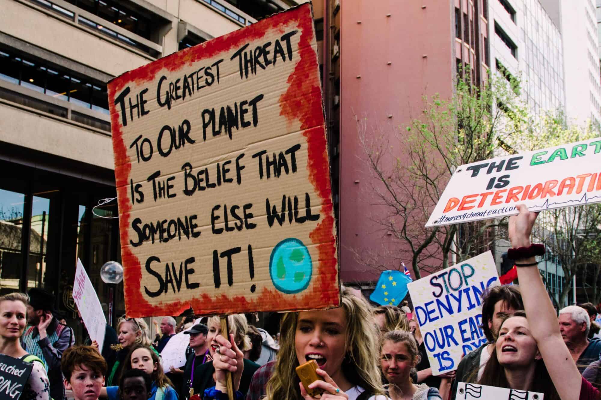 Climate protestors holding up a cardboard sign that reads "The greatest threat to our planet is the belief that someone else will save it"