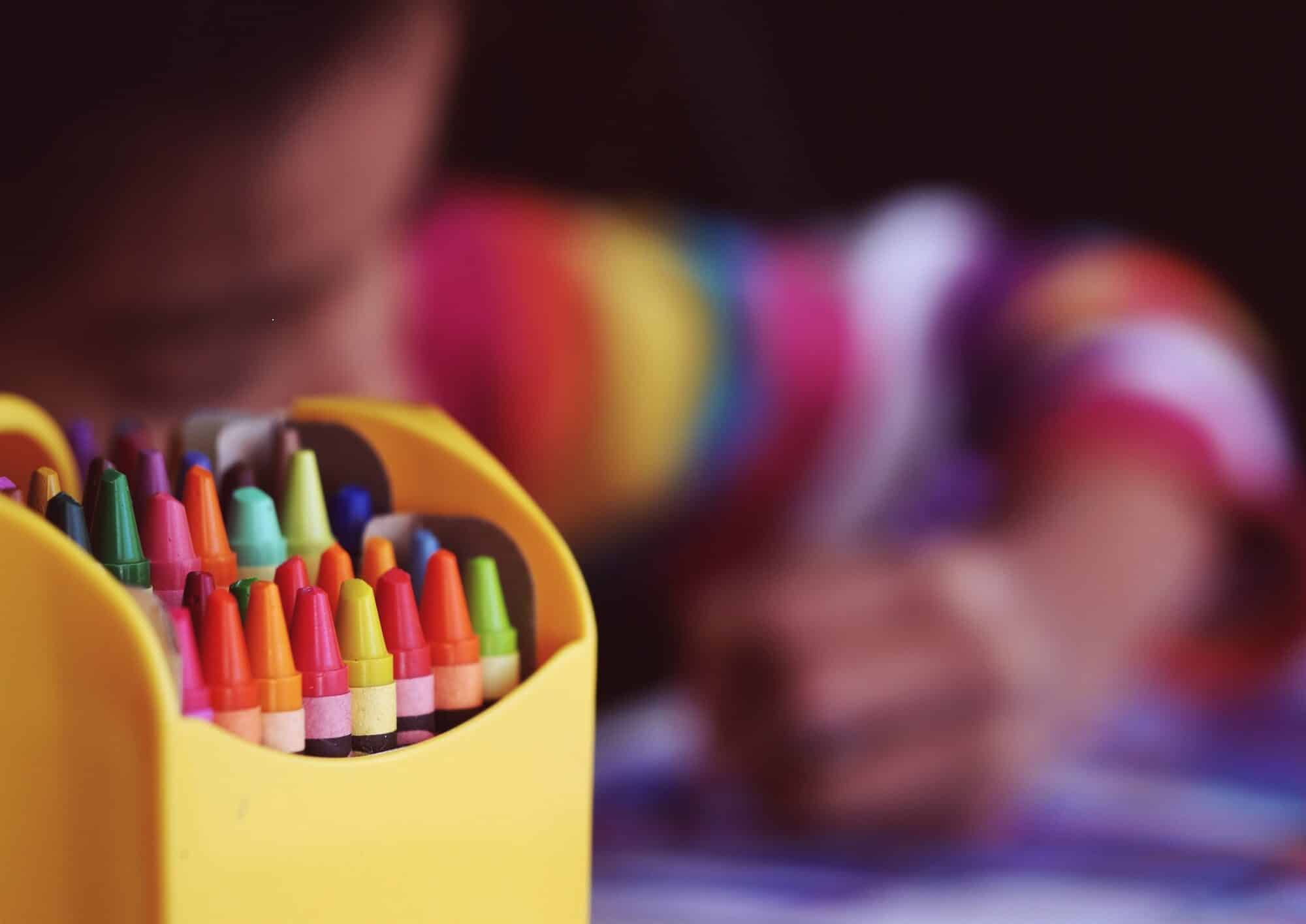 A pack of crayons in focus with a child in the background colouring.