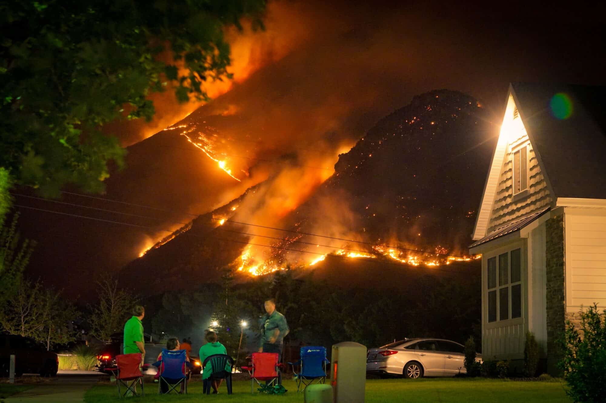 A family looks on from their driveway on lawnchairs as a wildfire approaches their home.