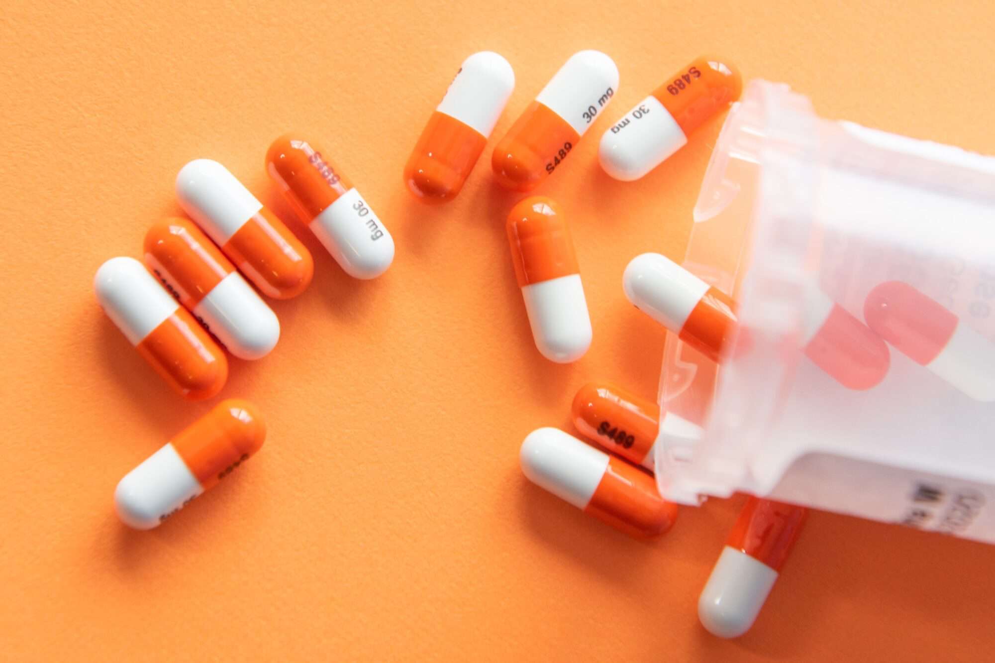 A clear pill bottle spills a number of orange and white capsules on an orange piece of paper.